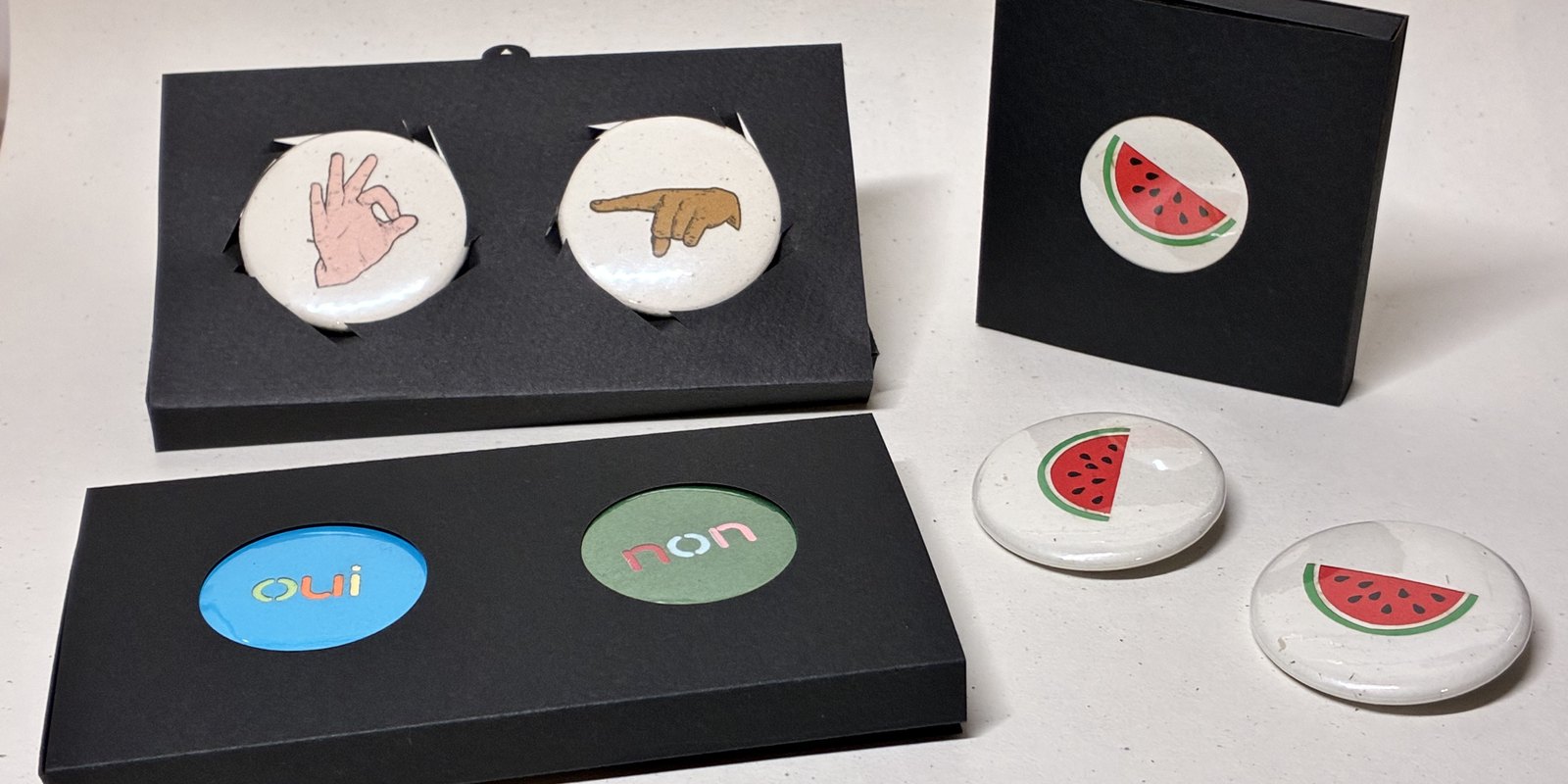 Badges Holy Sheet dans leurs boîtes noires à fenêtre découpée : pastèque pour la Palestine, main qui fait « OK », doigt qui pointe, « oui » sur bleu et « non » sur vert